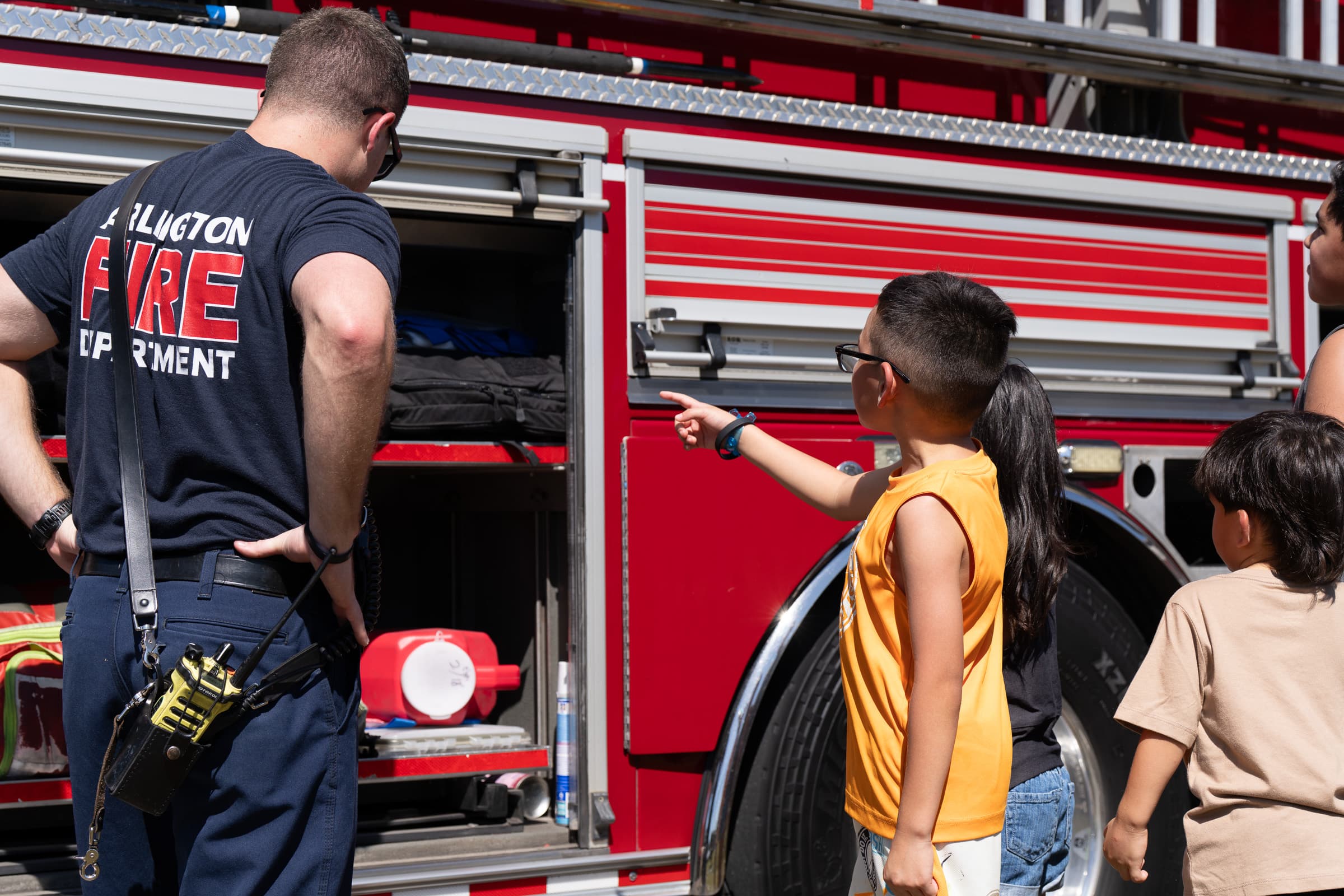 Firefighter showing children the tools and equipment stored in a red fire truck at a community event.