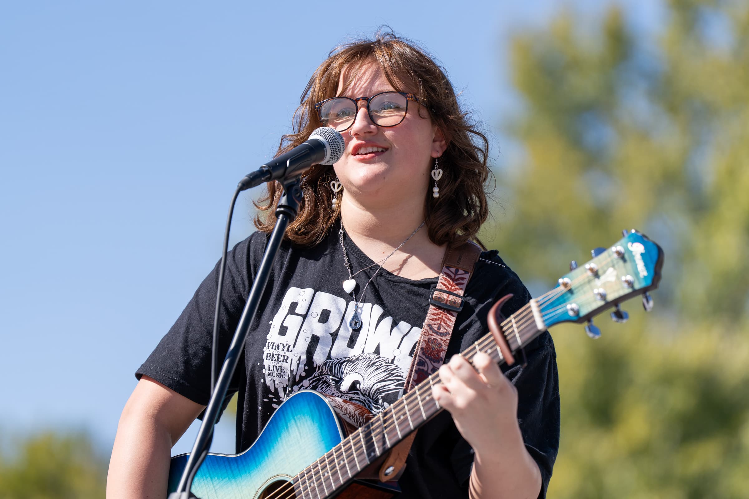 Young musician singing into a microphone while playing a blue acoustic-electric guitar at an outdoor event.
